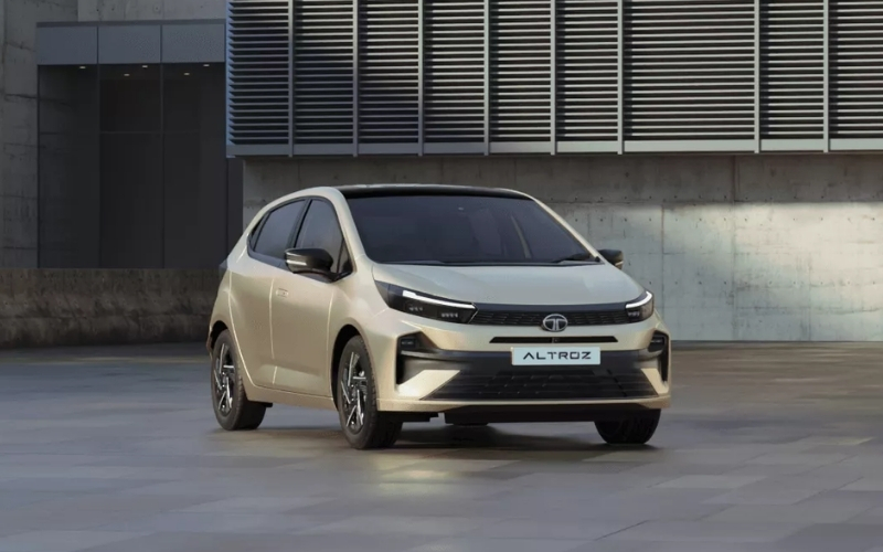 Front view of a 2025 Tata Altroz facelift parked in a modern, minimalistic urban setting. The car features a sleek, aerodynamic design with sharp LED headlights, a blacked-out grille, and sporty alloy wheels, finished in a light metallic color